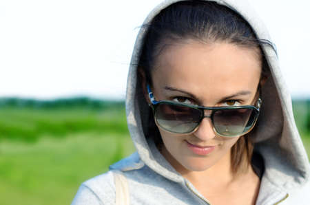 Woman in a hooded anorak standing in open countryside peering over her sunglasses at the camera with...