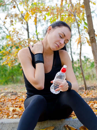 Woman athlete sitting on a curb in a park stretching her neck muscles and massaging them with her hand as she pauses for a drink of waterの写真素材