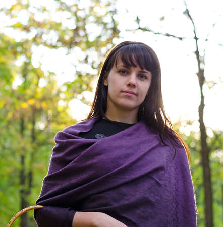 Attractive young woman wearing a purple shawl draped around her shoulder standing outdoors holding a wicker basket over her armの写真素材