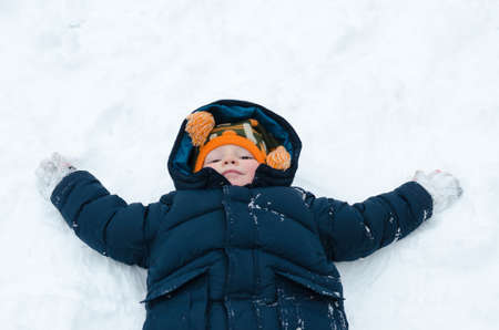 Little boy lying on his back in the snow in his winter jacket playing snow angels with his arms outspreadの写真素材