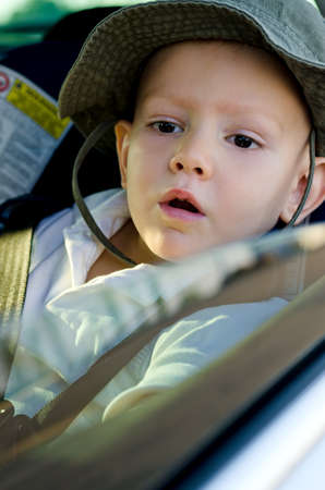 Adorable little boy wearing a hat waiting in a car strapped into a child safety seat and peering out...