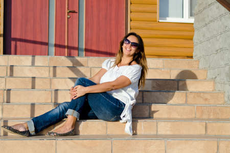 Attractive young happy smiling woman wearing sunglasses relaxing in the sun on the front steps of her houseの写真素材