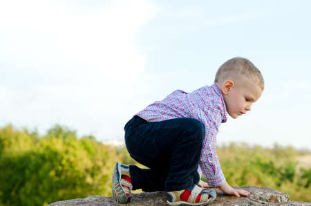 Little preschool boy exploring the countryside climbing onto a large rock, sideways view against cloudy blue skyの写真素材
