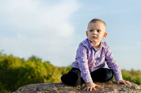 Little boy sitting on a rock outdoors on the sunshine enjoyingthe adventure of exploring the surrounding countrysideの写真素材
