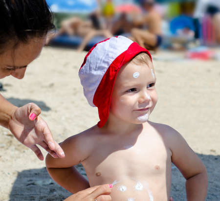 Cute little boy at the seaside wearing a colourful sunhat grinning happily as his mother draws a face on his tummy with sunscreenの写真素材