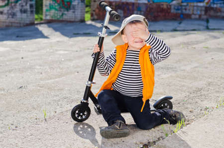 Bashful little boy sitting on the footboard of his scooter covering his eyes from the cameraの写真素材