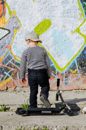 Little boy with his back to the viewer walking away from his scooter which is lying on the sidewalk towards a wll covered in colourful graffiti artの写真素材