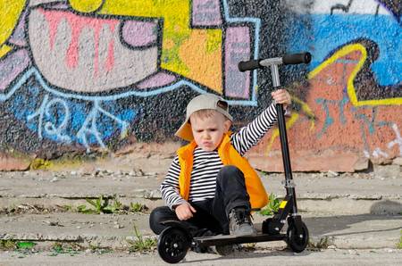 Tired little boy with a sulky expression sitting holding onto his scooter in front of a brightly coloured wall covered in graffitiの写真素材
