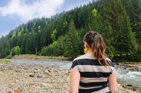 Woman standing with her back to the camera admiring a fast flowing mountain river in a forested valleyの写真素材