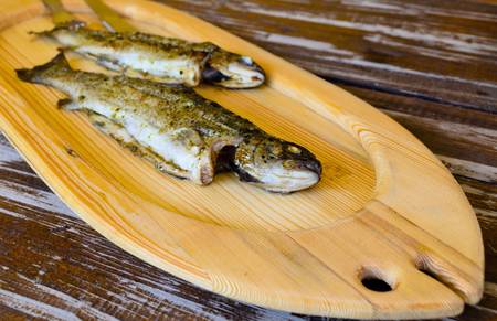 Two grilled fish resting on a fish shaped wooden board over an old wooden tableの写真素材