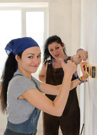 Pretty young woman giving a thumbs up of approval and success while renovating an apartment with a friend who is helping her align a spirit level on the wallの写真素材