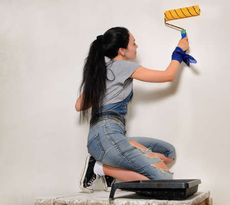 Young woman in trendy jeans with her brunette hair in a ponytail painting a wall with a roller during renovationsの写真素材