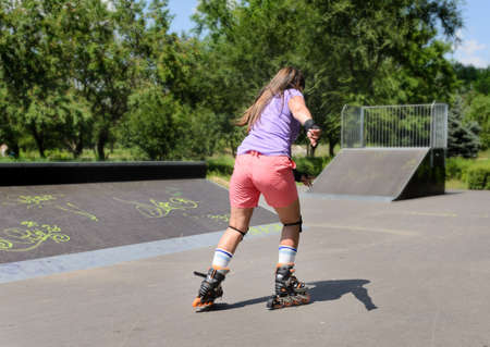 Young teenage girl rollerblading in a skate park speeding away from the camera toward distant cement...