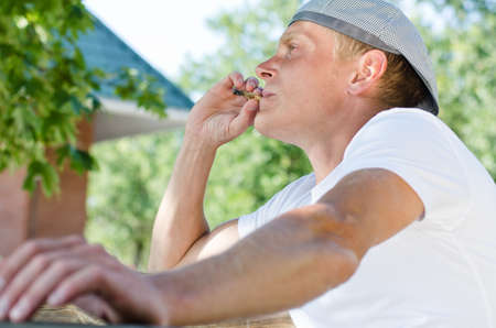 Trendy young man enjoying a smoke break sitting outdoors relaxing in the garden puffing on his cigarette , low angle close up viewの写真素材