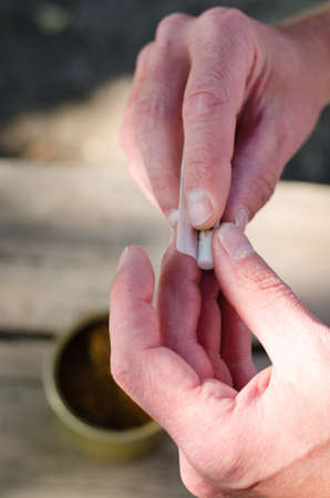 Man rolling himself a cigarette from loose tobacco blend using rollings, or adhesive paper covering sealed by licking, close up view of the handsの写真素材