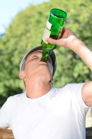 Low angle view of a young male alcoholic with a drinking problem gulping down alcohol from a bottle while sitting alone outdoorsの写真素材