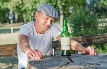 Mellow drunk sitting at an outdoor rustic wooden table in a park smiling at his bottle of alcoholの写真素材