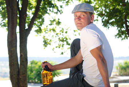 Man sitting relaxing in a rural landscape with a bottle of alcohol clutched in his hand turning to look at the camera with a serious expressionの写真素材