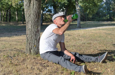 Alcoholic sitting drinking in a park swigging spirits directly from a bottle as he seeks to fill his craving and addictionの写真素材
