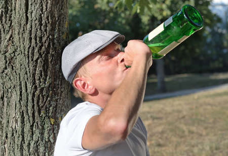 Drunk man with an alcohol problem standing up against the trunk of a tree in the sunshine gulping down spirits from a bottleの写真素材