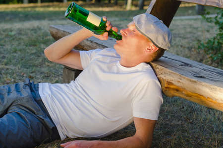 Jobless Caucasian alcoholic man drinking from a bottle of wine leaning on a bench in the parkの写真素材