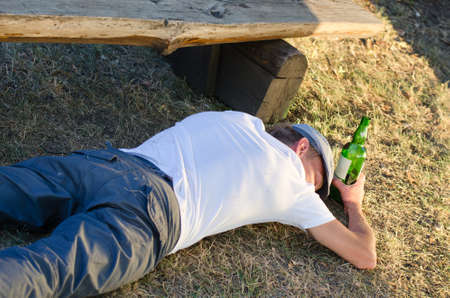 Drunk man holding a bottle of alcoholic beverage sleeping on the ground next to a bench in the park in summerの写真素材