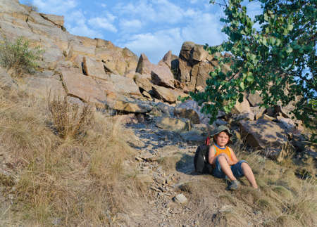 Cute little boy sitting on the ground and enjoying a warm day of summer at the mountains