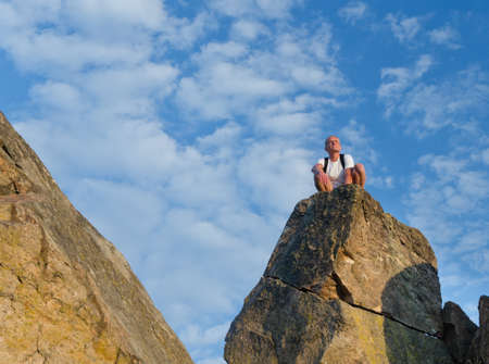 Man wearing a rucksack sitting on top of a rocky pinnacle on a mountain peak, against a blue summer skyの写真素材