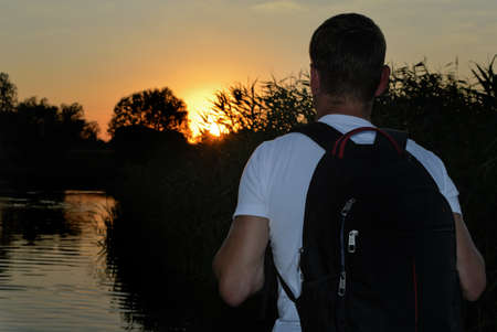 Backpacker standing watching the sunset overlooking a tranquil lake with tall reeds silhouetted against the colourful orange skyの写真素材