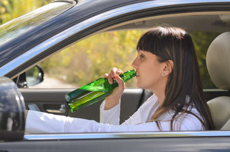 Alcoholic woman drinking and driving raising the bottle to her lips to take a swig as she steers the car, view through the side windowの写真素材