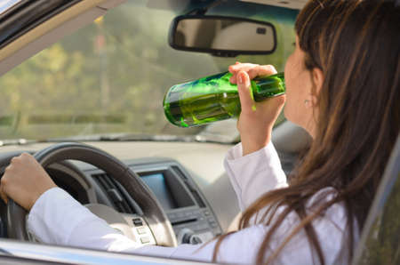 Woman drinking alcohol and driving raising the bottle to her lips to take a swig as she steers the...