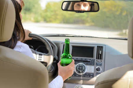 View inside a car from the back passenger seat of a woman driving holding a bottle of alcohol in one hand while steeringの写真素材