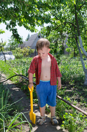 Young boy working in the vegetable garden standing on the path with an open shirt and his spade contemplating the plants on the side as he helps out during the summer vacationの写真素材