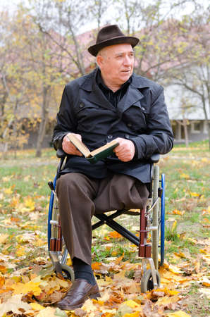 Handicapped elderly gentleman in a wheelchair sitting outdoors enjoying the autumn sunshine with his bookの写真素材
