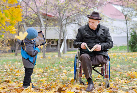 Disabled grandfather sitting in a wheelchair watching his young grandson playing with a tablet computer as the two enjoy a relaxing day in an autumn park togetherの写真素材