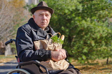 Senior handicapped man doing his grocery shopping sitting in the road in his wheelchair clutching a large bag of food and looking at the cameraの写真素材
