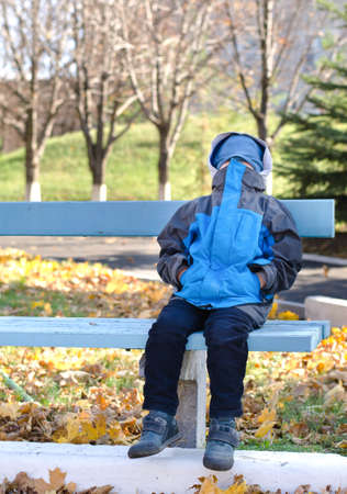 Young boy sitting into park on a bench with his face covered as he plays at being scary, in defiance as he sulks, or trying to be secretive and mysteriousの写真素材