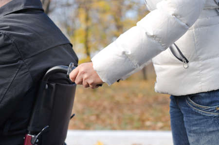 Close up view of the hands of a woman pushing a disabled man in a wheelchair along a rural street in...