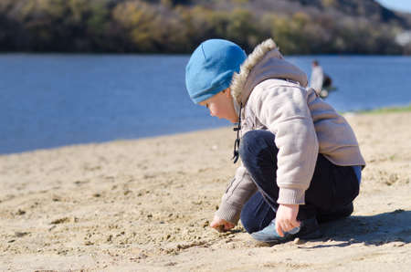 Cute little boy playing on a sandy beach crouching down in his beanie cap and jeans drawing in the sand at the edge of the waterの写真素材