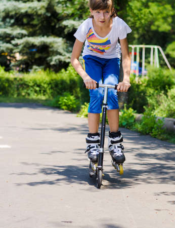 Teenage girl having fun on a scooter as she tries to balance in her roller blades on the footboard and propel herself along the tarmac in a rural parkの写真素材