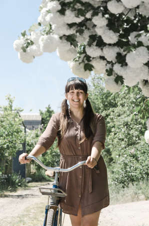 Beautiful woman with a bicycle smiling as she admires bunches of pretty white spring blossom on a tree in a rural gardenの写真素材