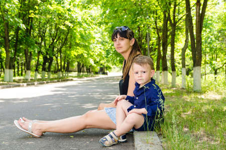 Mother and her cute little son waiting in a tree-lined rural street sitting together on the curb as they wait for someone to arriveの写真素材