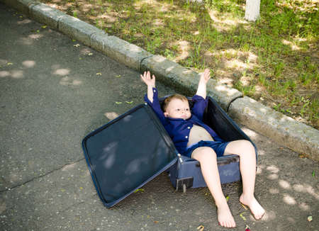 Excited little boy lying inside a suitcase cheering and raising his arms in the air as he waits for...