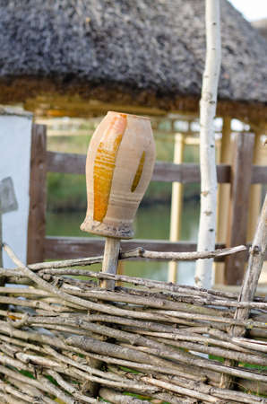 Rustic clay pot painted with yellow stripes upended on a pole outdoors in the sunshine behind a simple woven wood fenceの写真素材