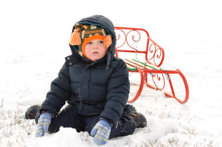 Small boy wrapped up warmly against the winter cold kneeling in the snow with a bright orange metal...