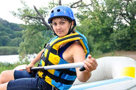 Active attractive middle-aged woman going river rafting sitting on a inflatable raft in a helmet and life jacket holding a paddleの写真素材