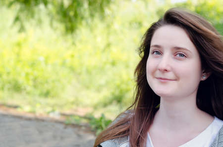 Beautiful young woman daydreaming in a lush green park standing with a serene expression and gentle...