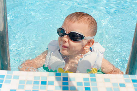 Laughing little boy climbing out of a swimming pool on the aluminium stepladder wearing goggles and...