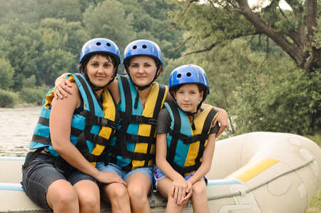 Two women and a child going white water rafting sitting on the edge of an inflatable raft in their...