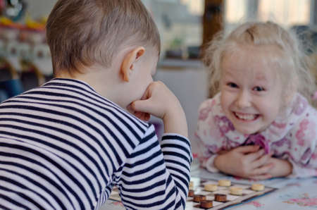 Cute little girl grinning mischievously at her brother as they sit at a table together playing a game of checkers or draughtsの写真素材